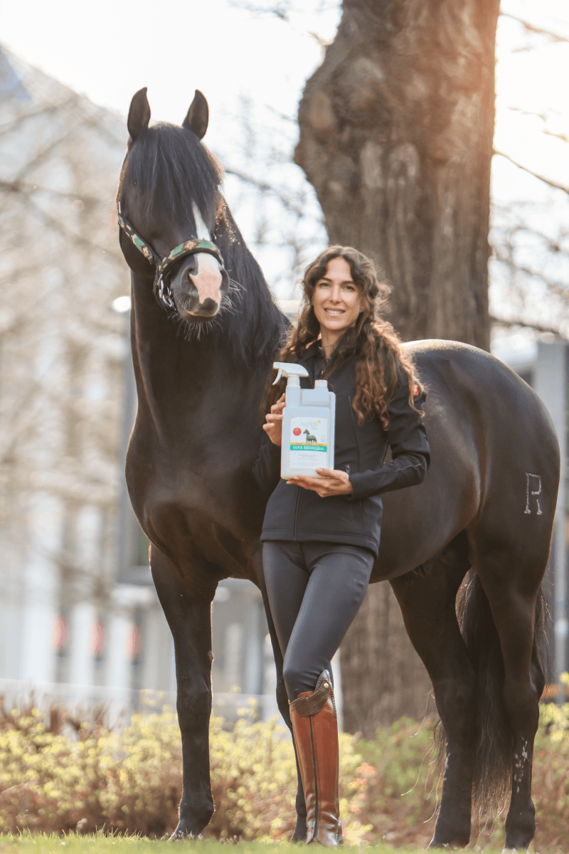 Pferdetrainerin und Schauspielerin Kenzie Dysli steht vor ihrem Pferd Fideo und hält das Produkt Josera Help Eukabronchial in der Hand